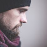 a side profile of a bearded man with a wooly hat on and a red scarf who looks sad